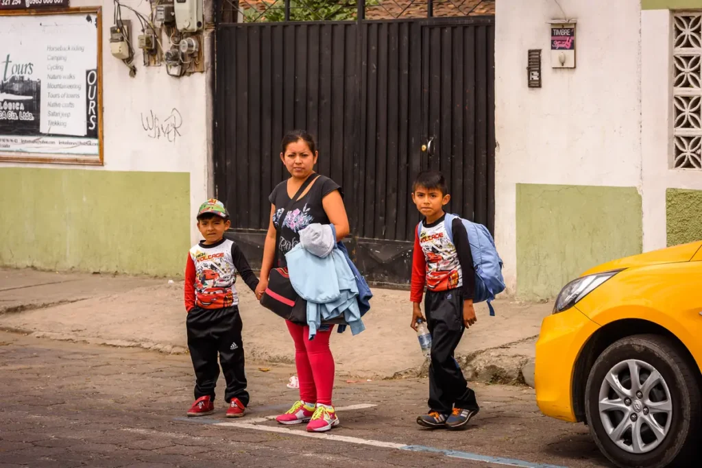 A woman walks two schoolboys with backpacks across a street in a small town.