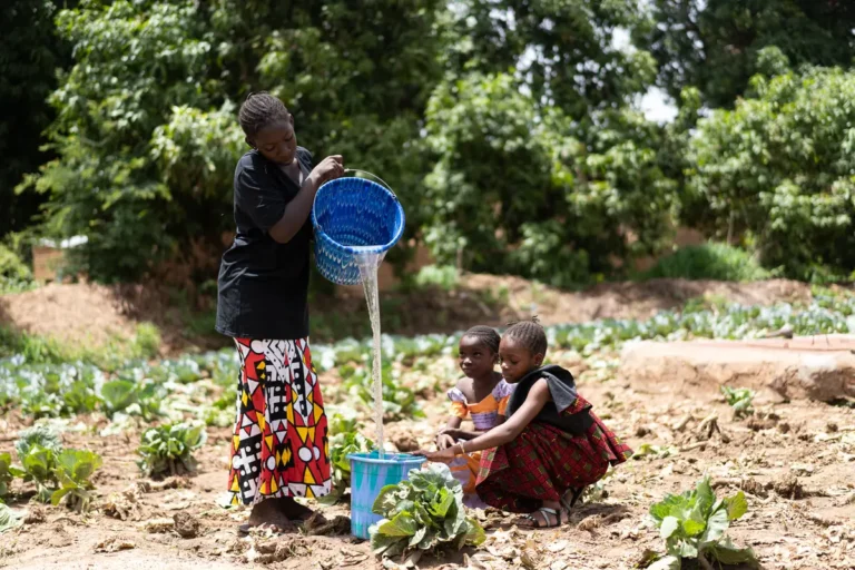 A young woman pours water into a bucket in a vegetable garden while two girls tend the plants.
