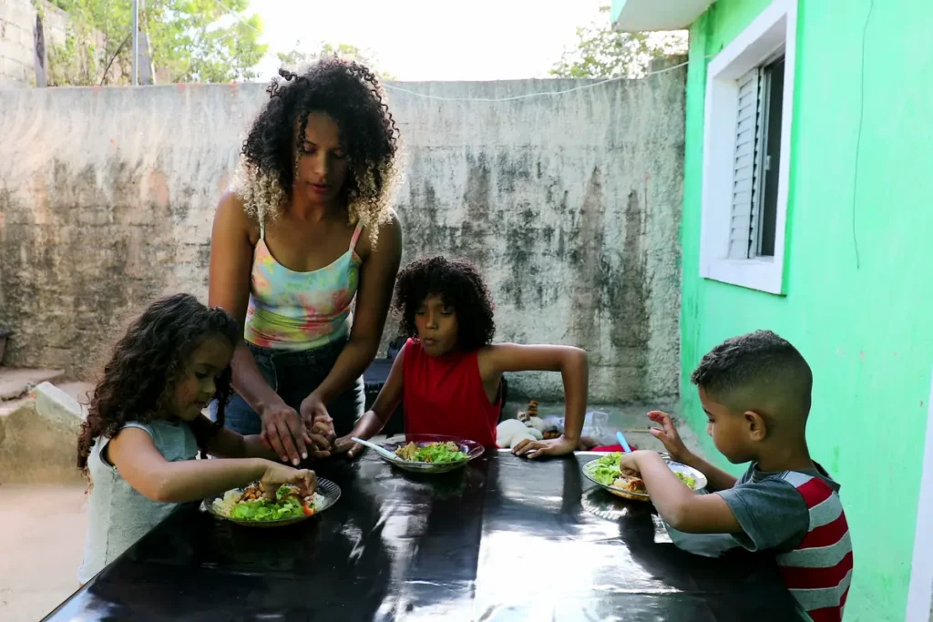 A woman serves a meal to three children at an outdoor table in a modest courtyard.